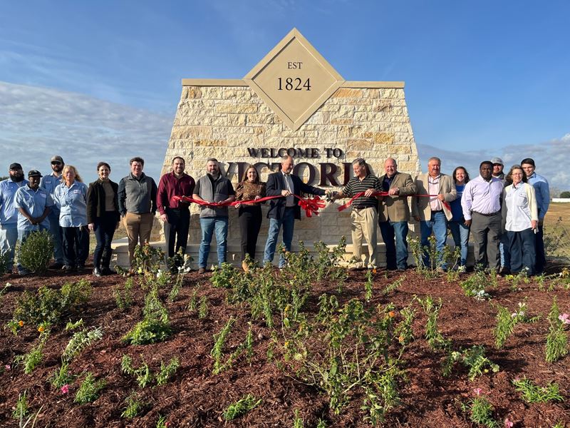 Large group of people stand in front of a welcome sign and cut a red ribbon