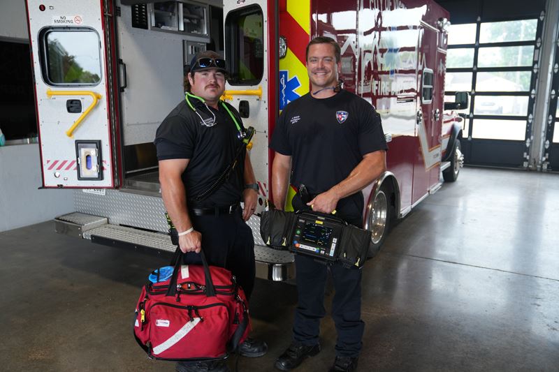 Two firefighter EMTs standing near an ambulance holding equipment kits