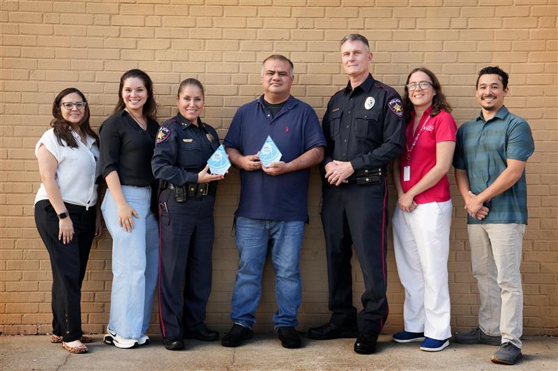 Group photo of 7 people, 2 in police uniform. Two of the people hold trophies.