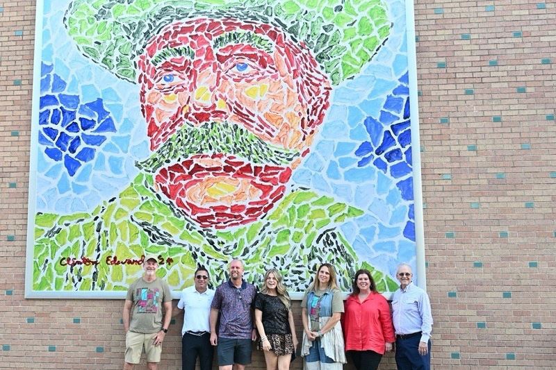7 people pose in front of a mural in downtown Victoria of Martin De Leon painted in a mosaic style