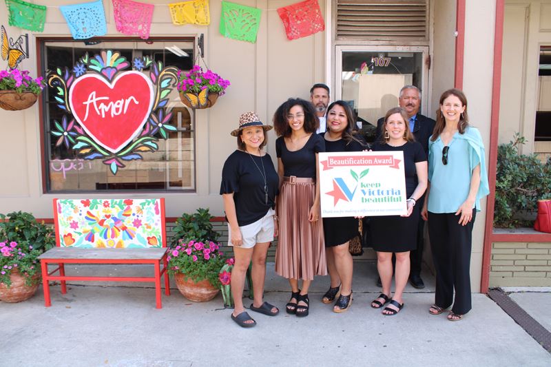 Group photo with KVB award sign at storefront with window painting, a colorful bench and decorations