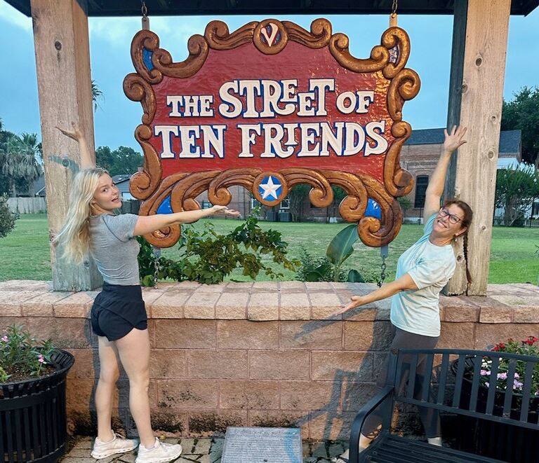 Two women pose gesturing to the Street of Ten Friends sign Opens in new window