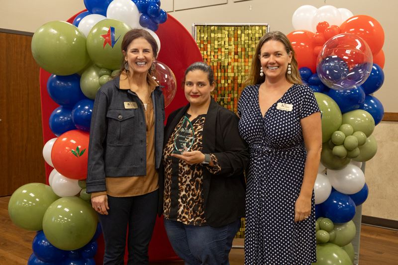 Three people in front of a balloon arch. The woman in the middle holds a trophy