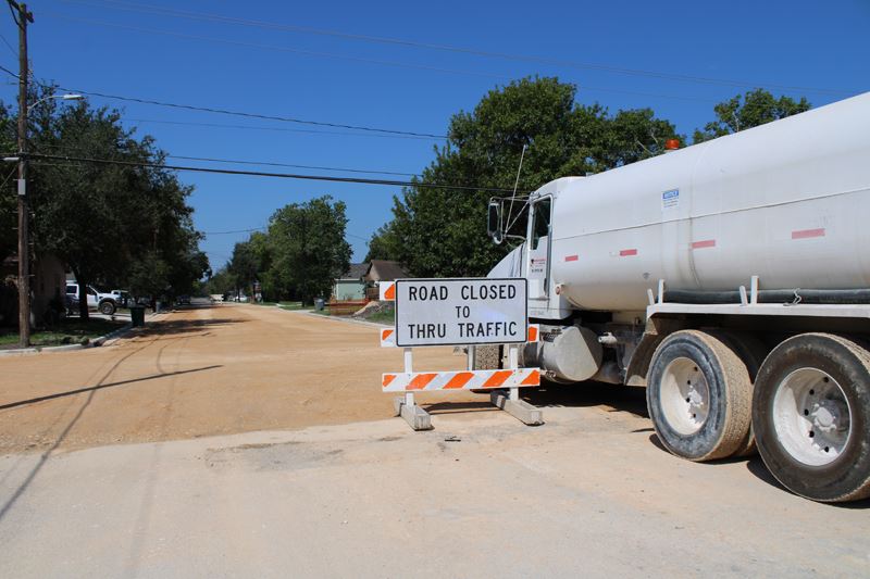A truck and a barricade sit at the edge of a street with no pavement but a layer of brown dirt.